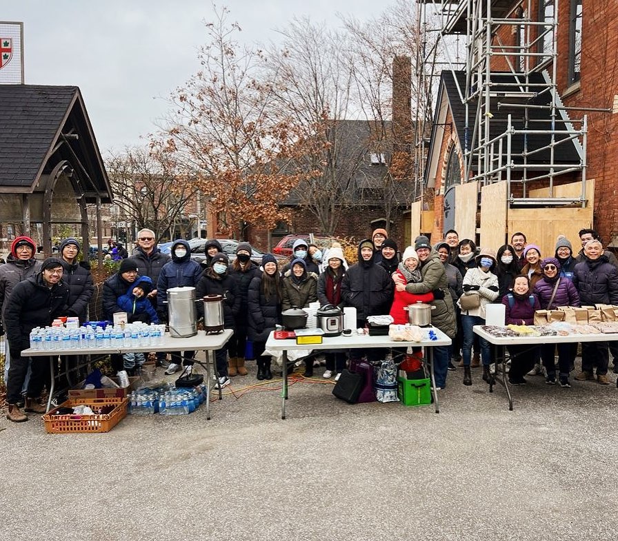 A large group of people standing behind tables offering food and drinks to people. In the background is Epiphany St Mark church.