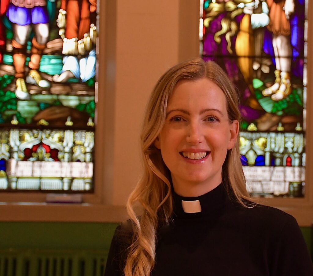 Closeup photo of curate Hannah Johnson wearing black with a clerical collarwith stained glass in the background